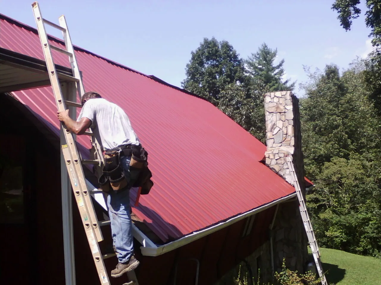Roofer on a ladder beside a red metal roof and stone chimney — Soffit & Fascia Repair by Roofing Brothers in Okahumpka